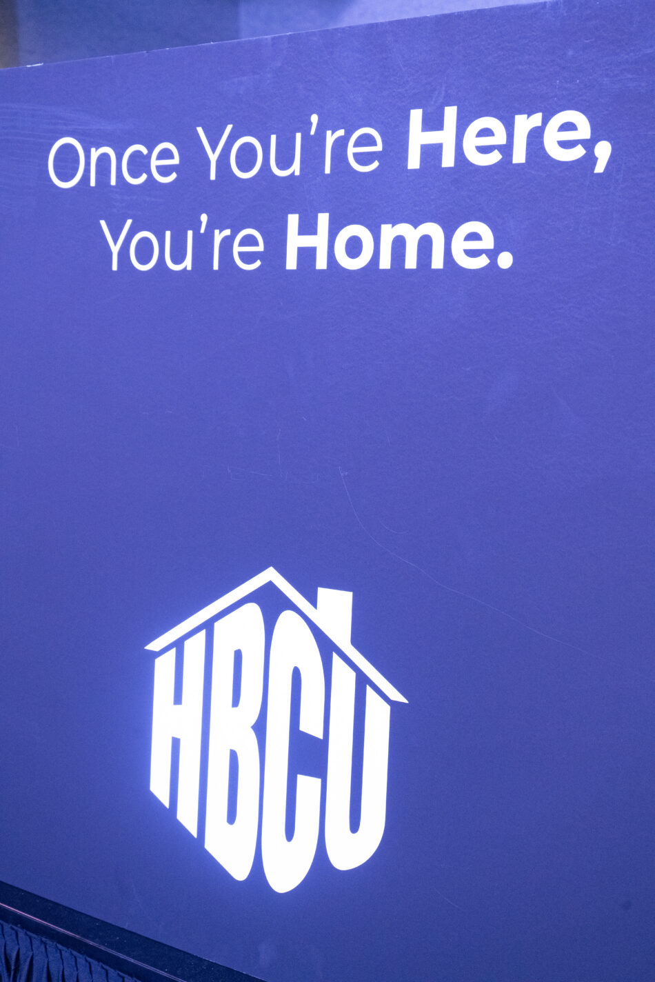 Blue sign with white text reading "Once You're Here, You're Home" above a house-shaped HBCU logo, symbolizing community and belonging.