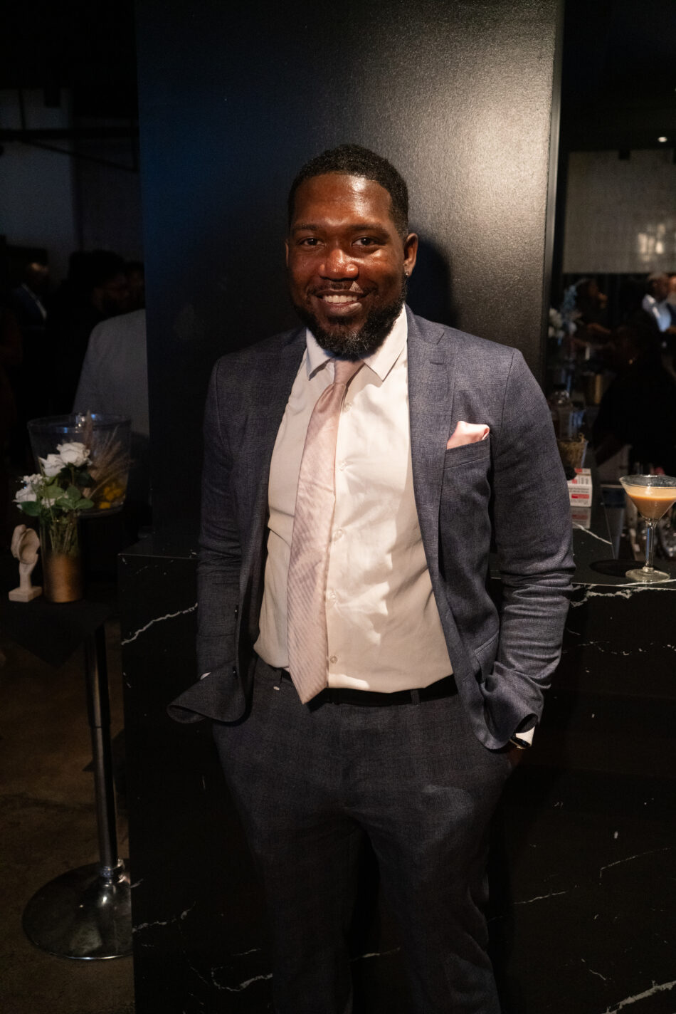 Smiling man in a sleek navy suit with a pink tie and pocket square, standing confidently at a professional networking event.