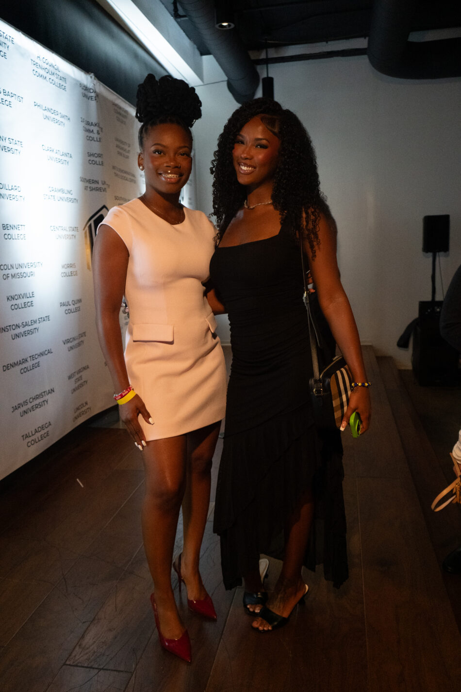 Two smiling women, one in a light pink dress and red heels, the other in a black dress, stand at an event.